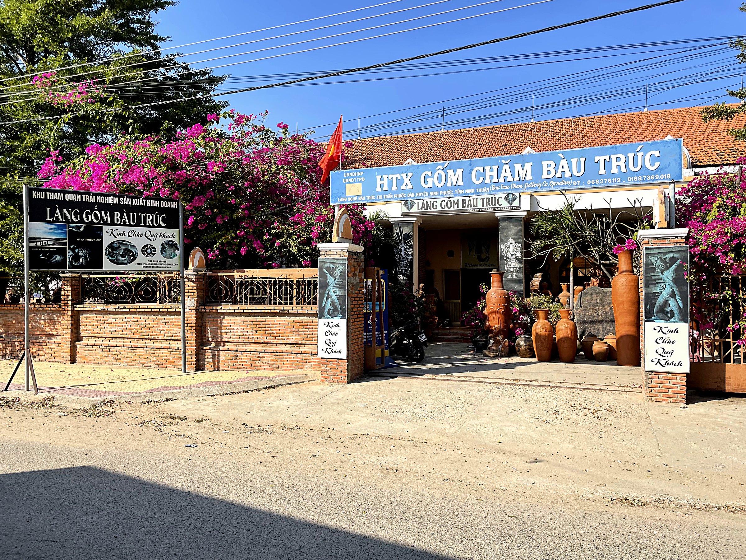 Cổng Hợp tác xã Gốm Chăm Bàu Trúc nhìn từ đường làng, hoa giấy hồng, bình gốm lớn trước cửa