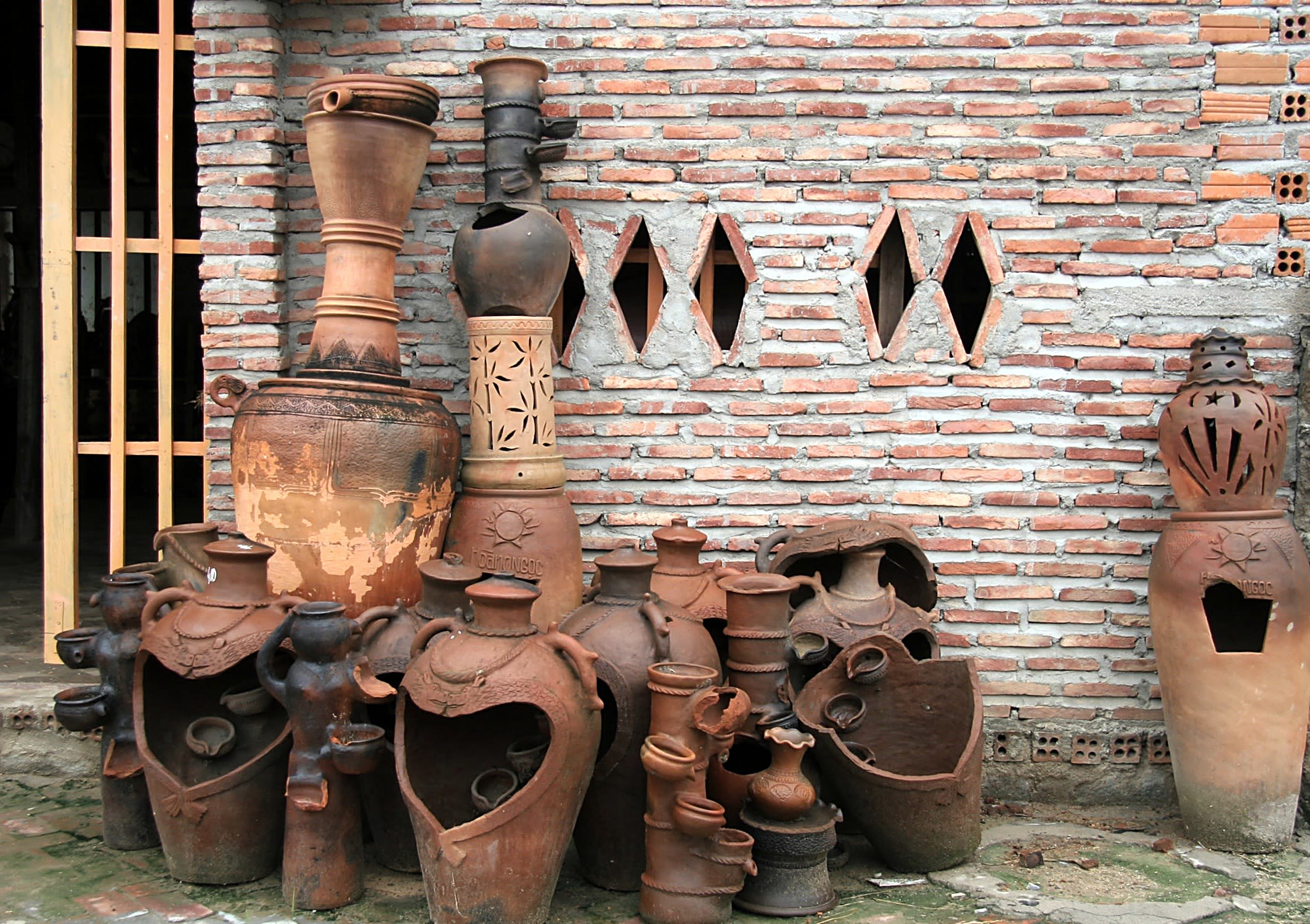 Bình và chậu đất nung đứng sát bức tường gạch, mặt ngoài loang lổ, vài chiếc khoét lỗ hình trái tim