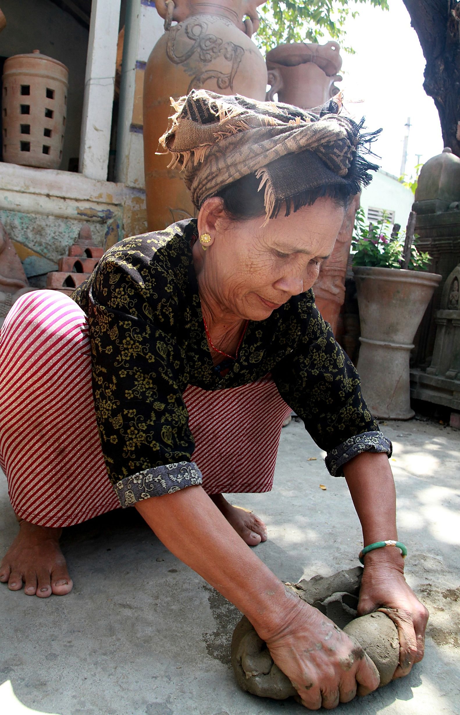 Người thợ nữ lớn tuổi ngồi xổm, hai tay nhồi khối đất sét trên nền sân